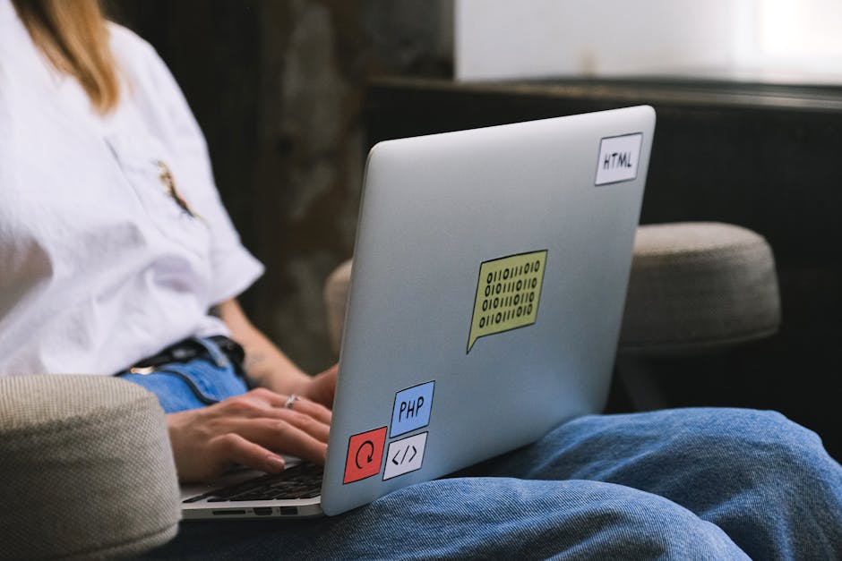 A person sitting on a beige upholstered couch outdoors, working on a silver laptop that is decorated with various colorful stickers including labels for HTML, PHP, a red power button, and a coding sym