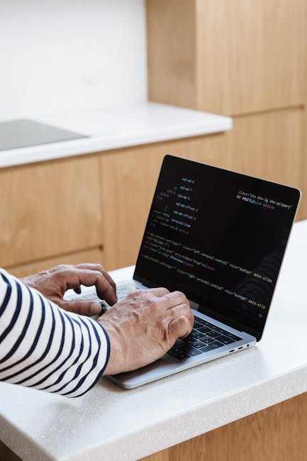 A person sitting on a beige upholstered couch outdoors, working on a silver laptop that is decorated with various colorful stickers including labels for HTML, PHP, a red power button, and a coding sym