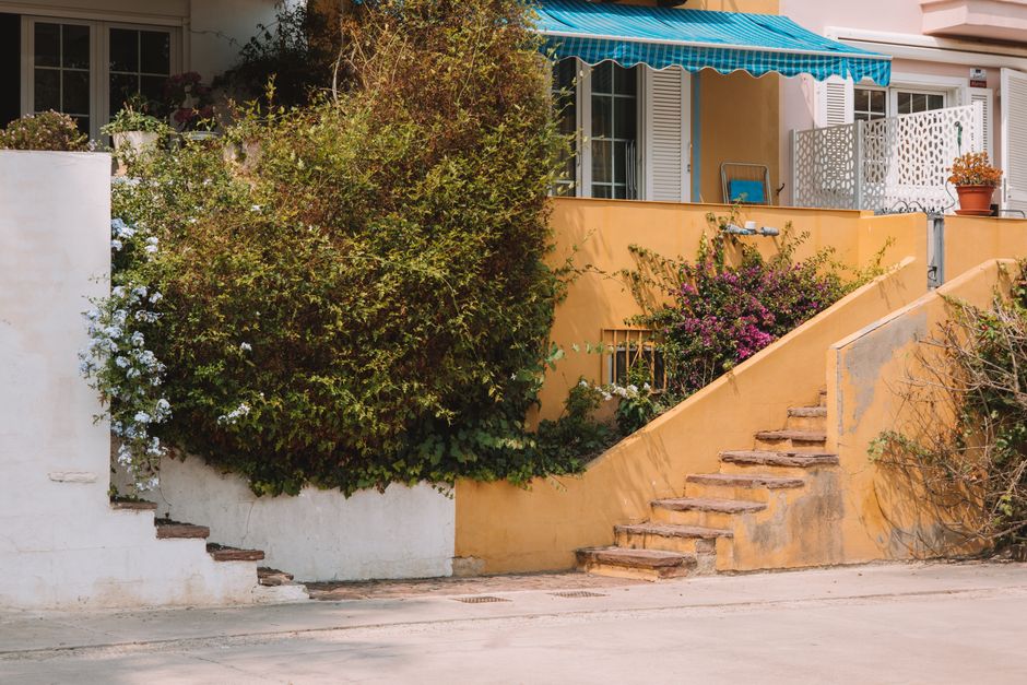 A terraced house with pastel yellow exterior walls and white window shutters features outdoor garden areas accessed via a short flight of terracotta-colored stairs with visible signs of weathering. Th